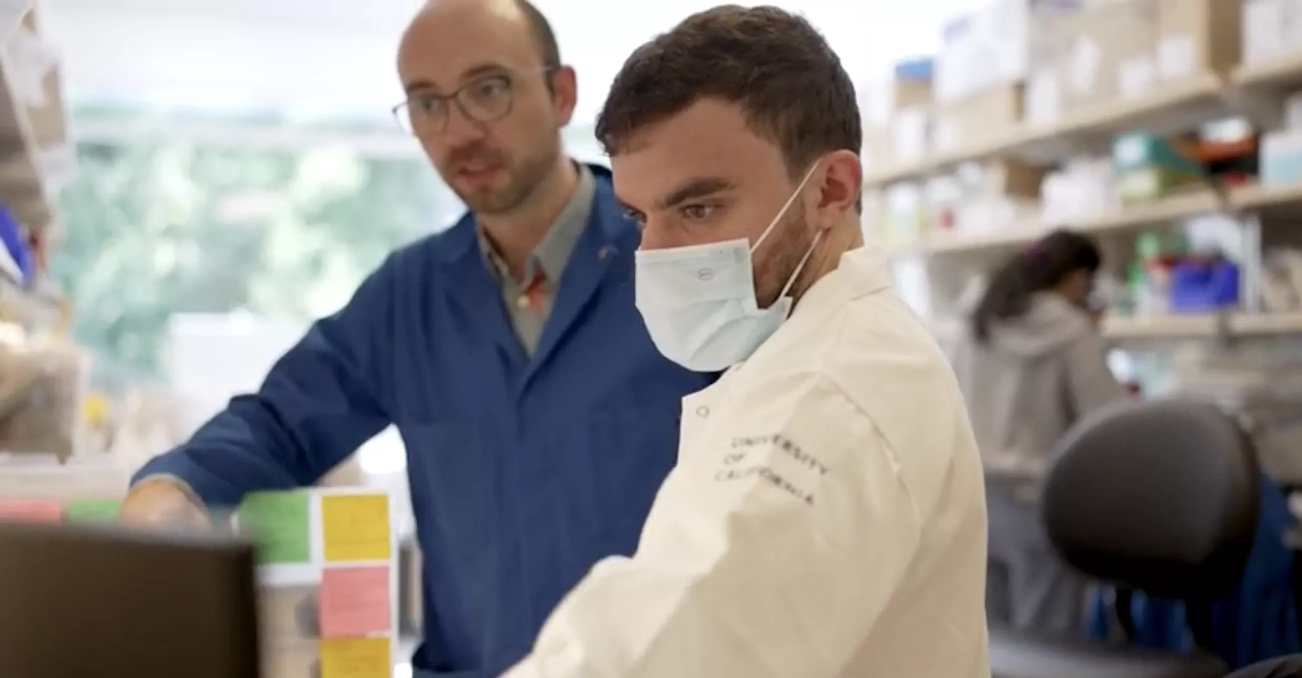 Man with short brown hair, lab coat and face mask gazes at a screen with another man in blue lab coat, glasses and very short brown hair in background blurry lab setting.