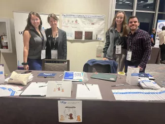 Two women in grey suits and a woman and man stand on opposite sides of a scientific poster smiling while at a booth with flyers, pens, and other marketing items laid out