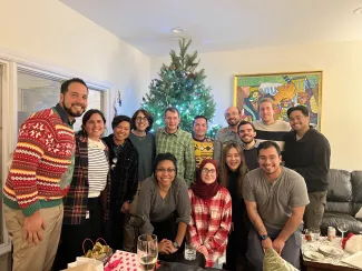 A group poses in front of a christmas tree with lights dressed in ugly sweaters and smiling in a living room setting